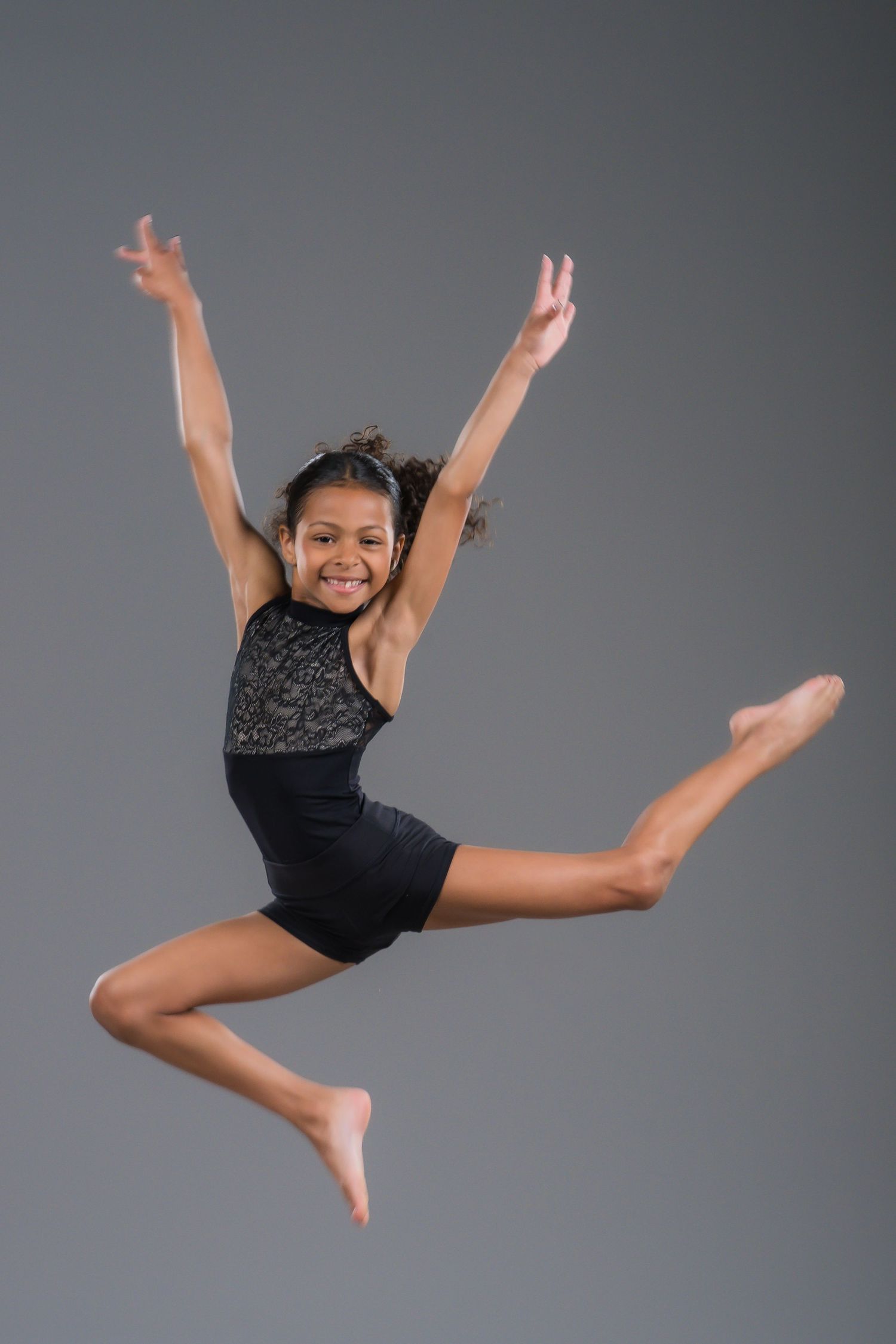 A smiling young girl in a black leotard performs a joyful leap with arms raised in a dance studio.