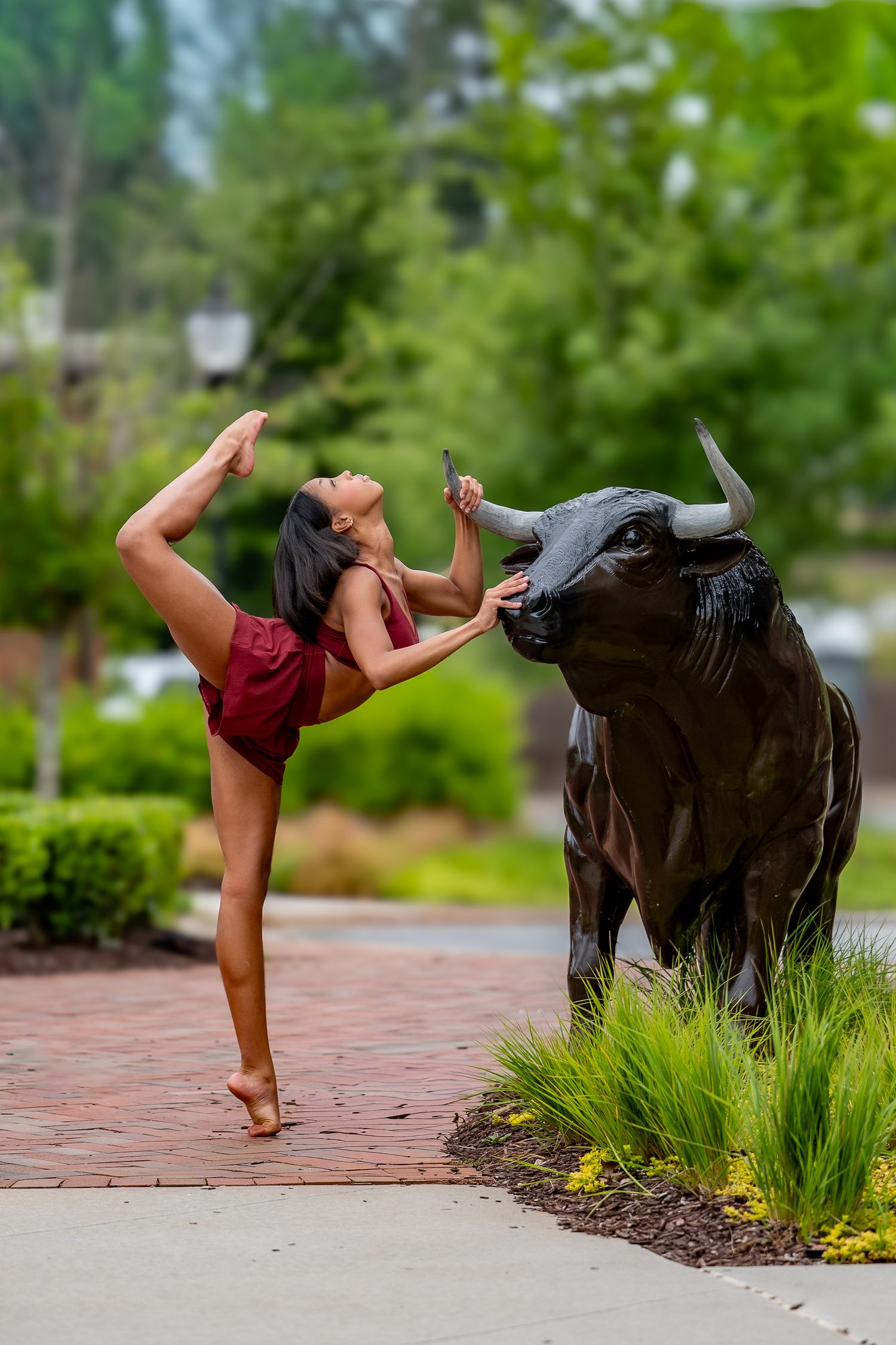 A dancer in red performs a standing split beside a bronze bull statue in a lush park.