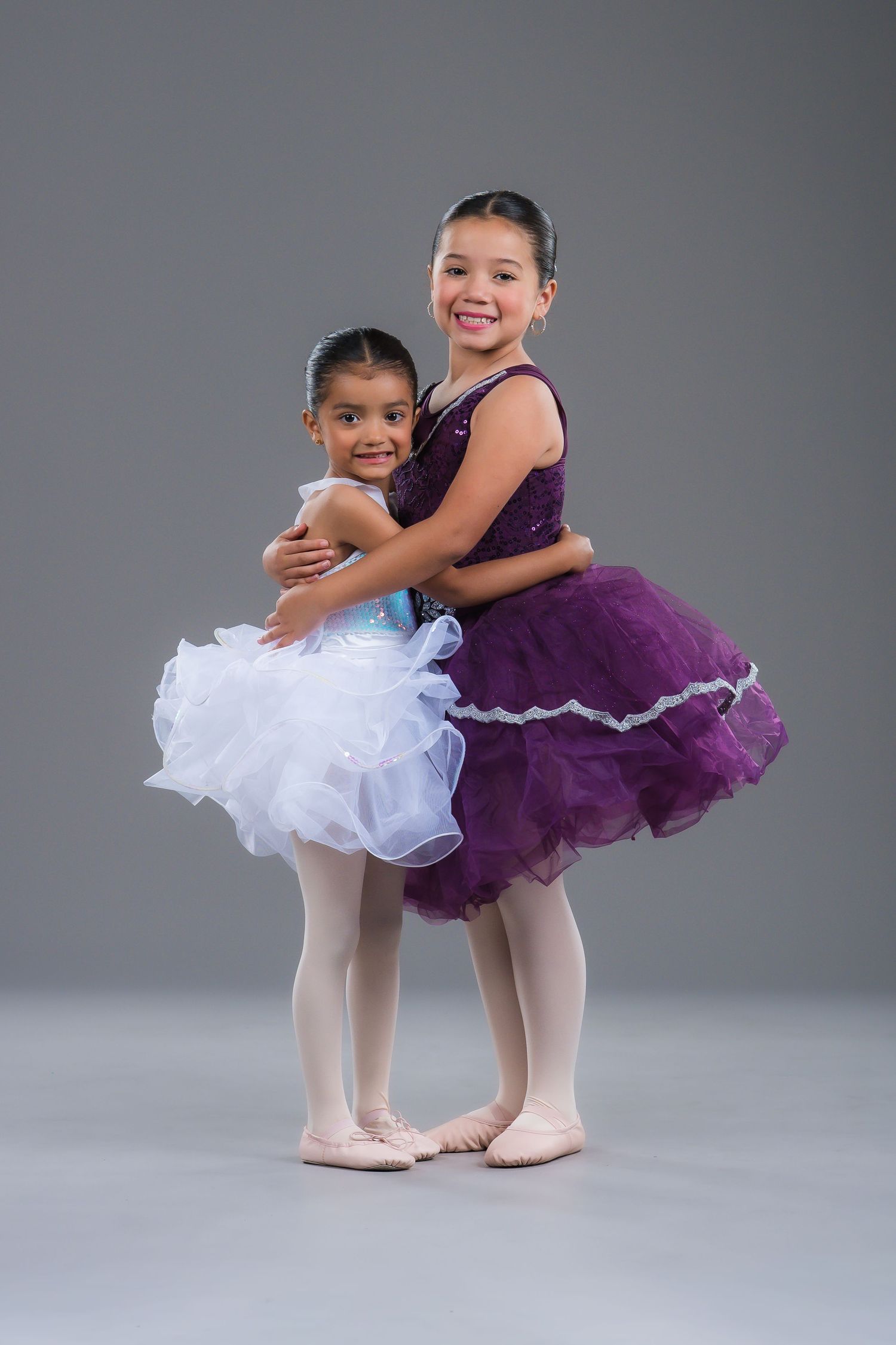 Two young ballerinas in purple and white tutus embrace and smile in a gray studio setting.