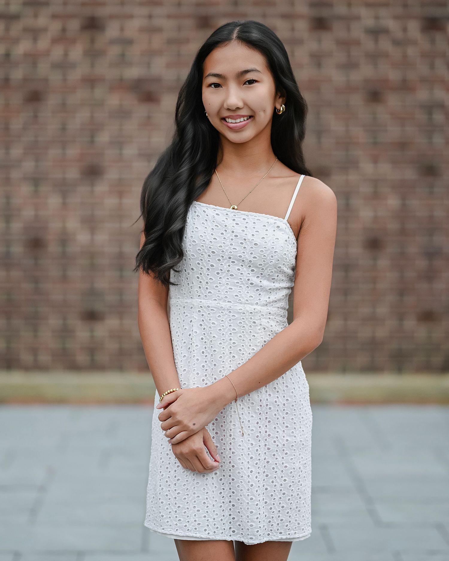 Smiling Asian teenage girl in white sundress posing for senior portrait in front of brick wall.