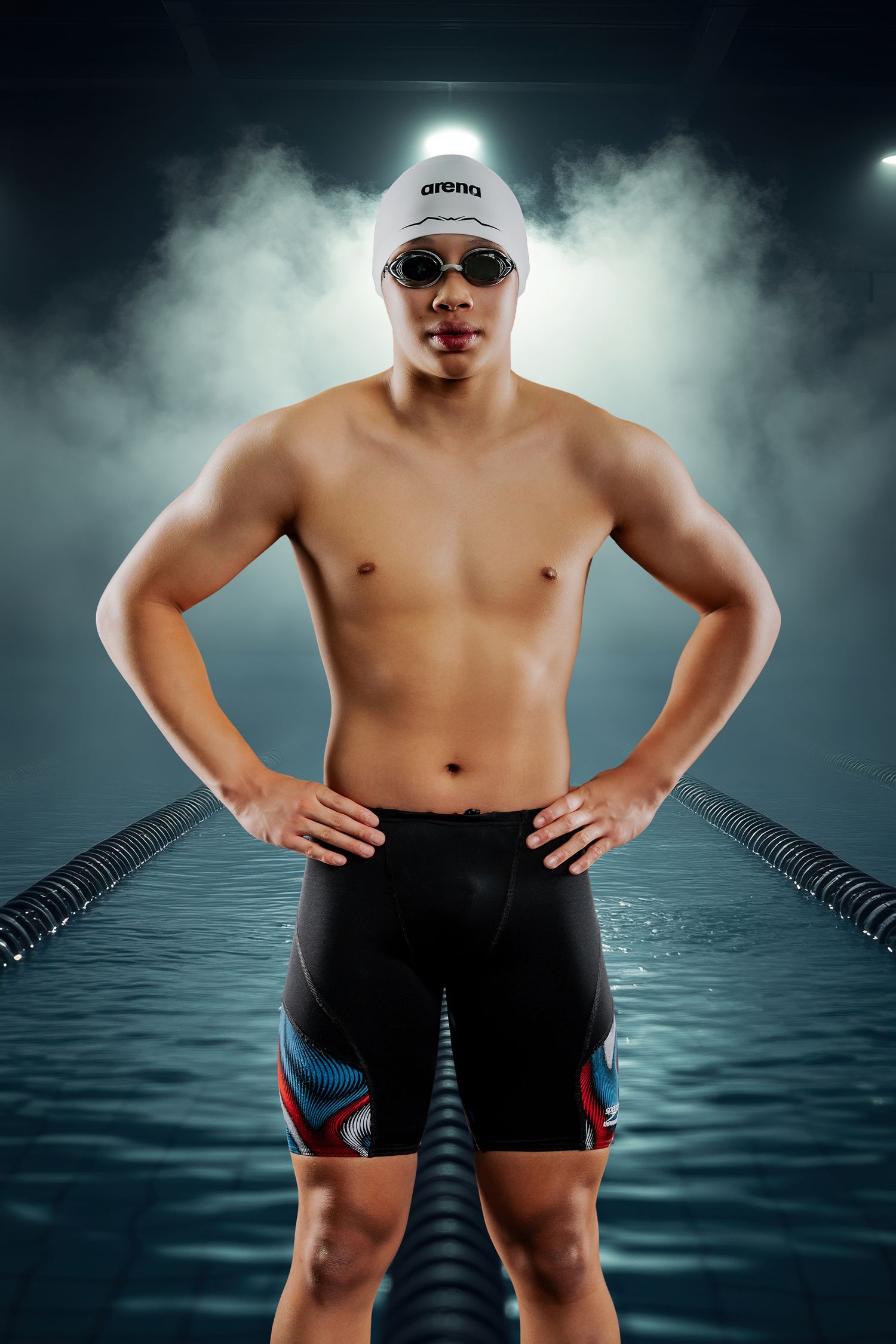 Male swimmer in Arena cap and goggles stands confidently at pool lane, dramatic lighting with misty atmosphere.