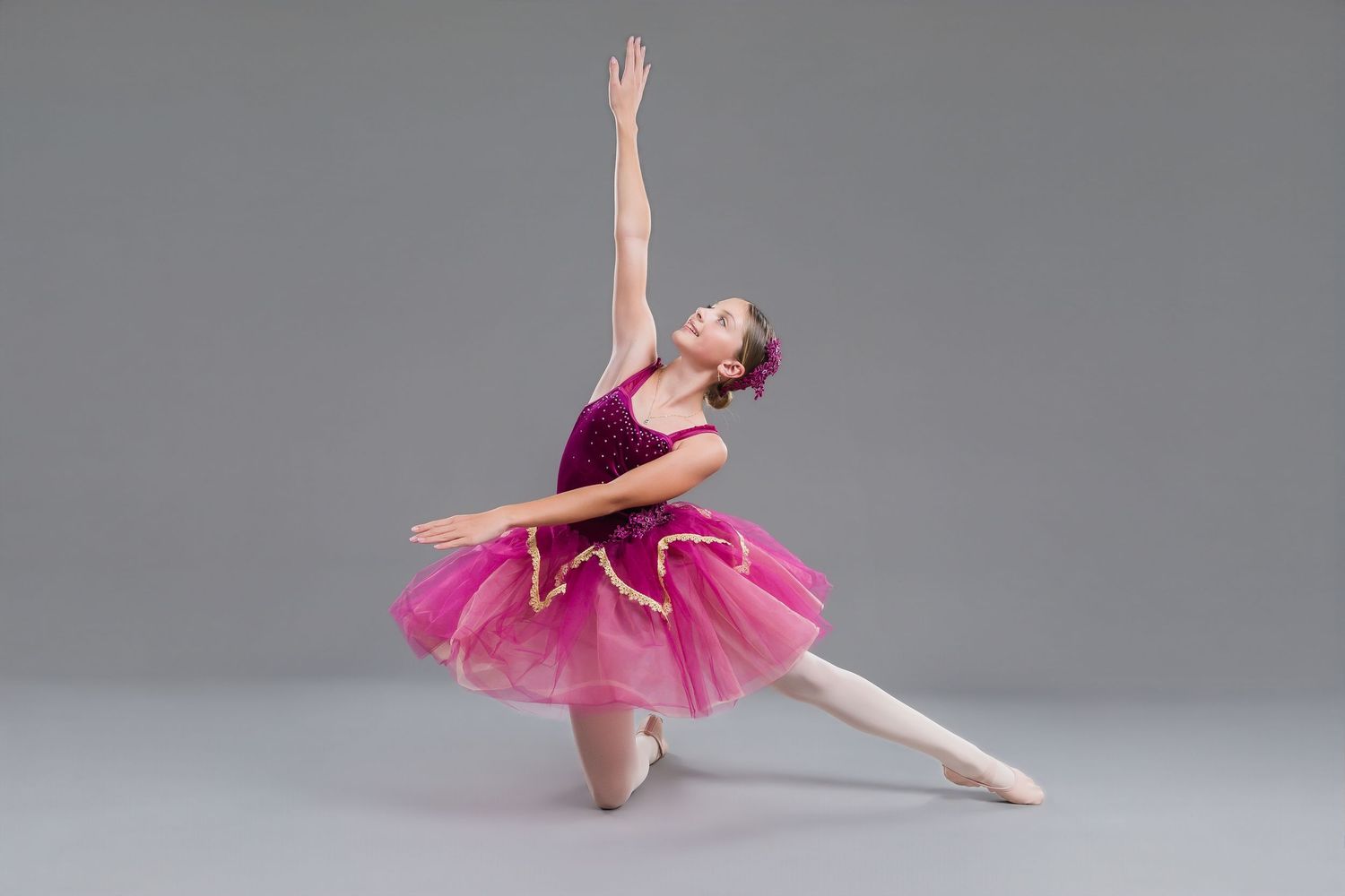 A young ballerina in a pink and purple tutu performs an arabesque pose in a studio.