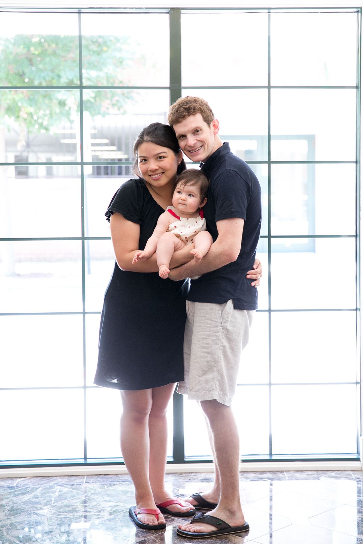 Happy interracial couple in matching black tops holding their baby indoors near large window.