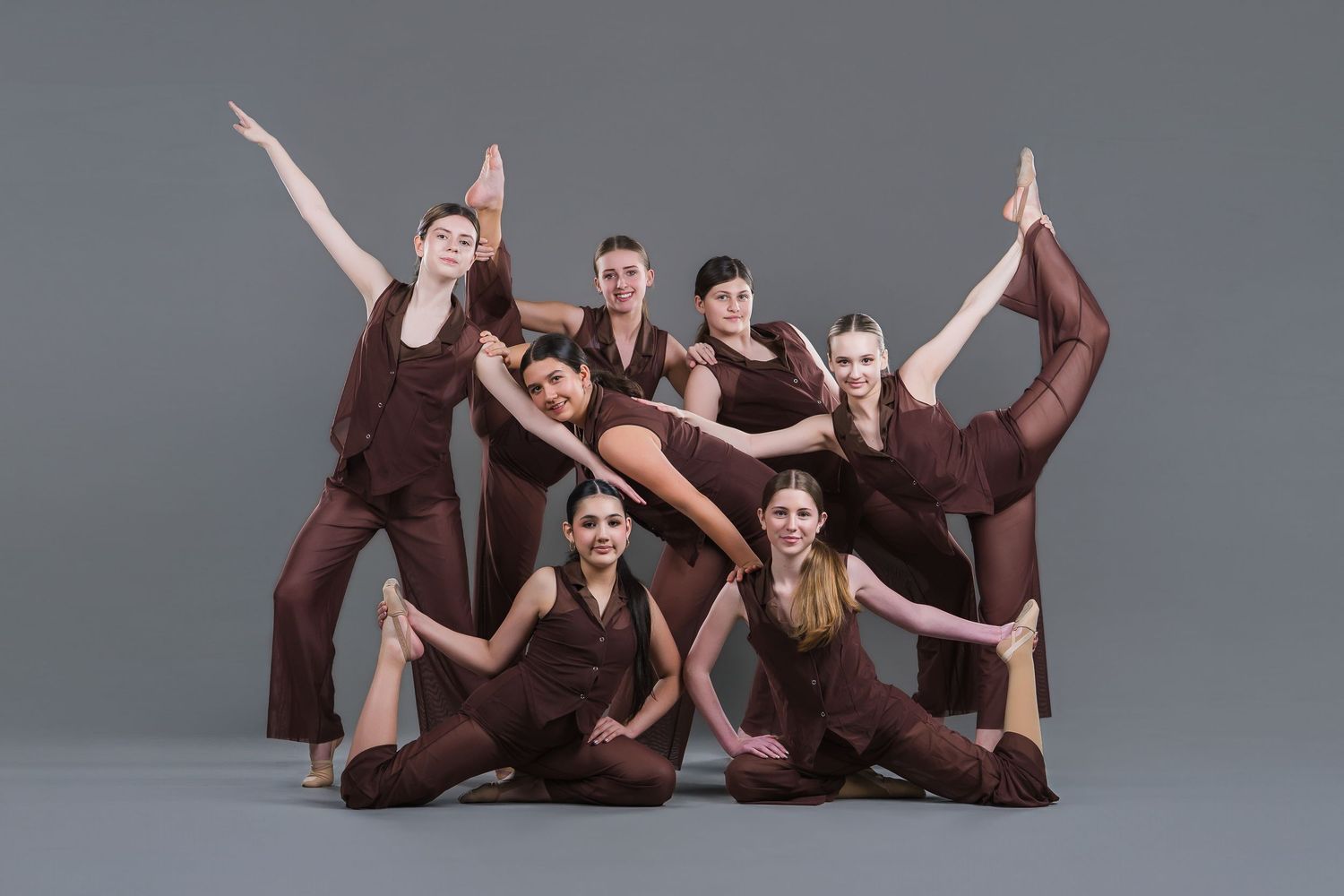 A group of seven contemporary dancers in brown costumes perform a dynamic studio group pose.