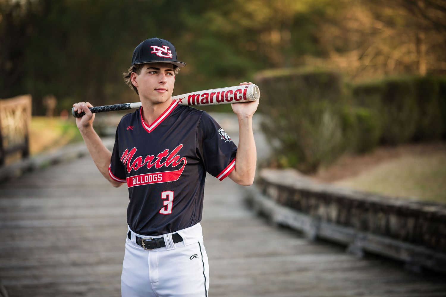 Baseball player number 3 in North uniform holds Marucci bat on wooden bridge.