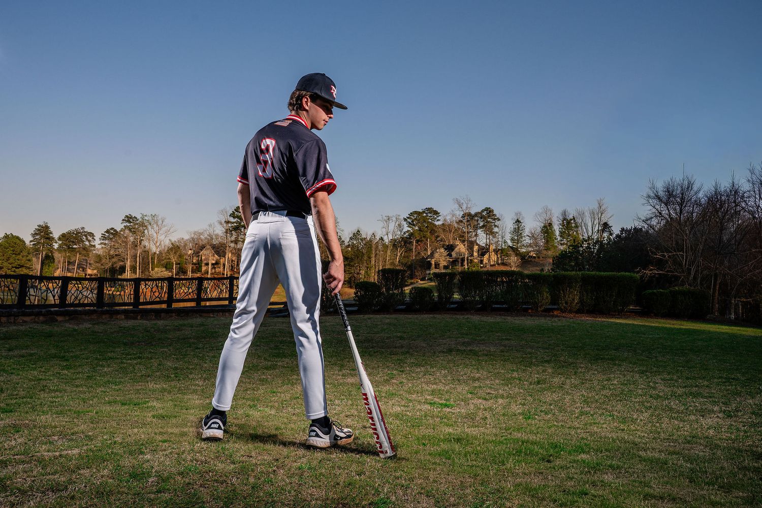 Baseball player in uniform stands with bat on sunny green field near wooden bridge.