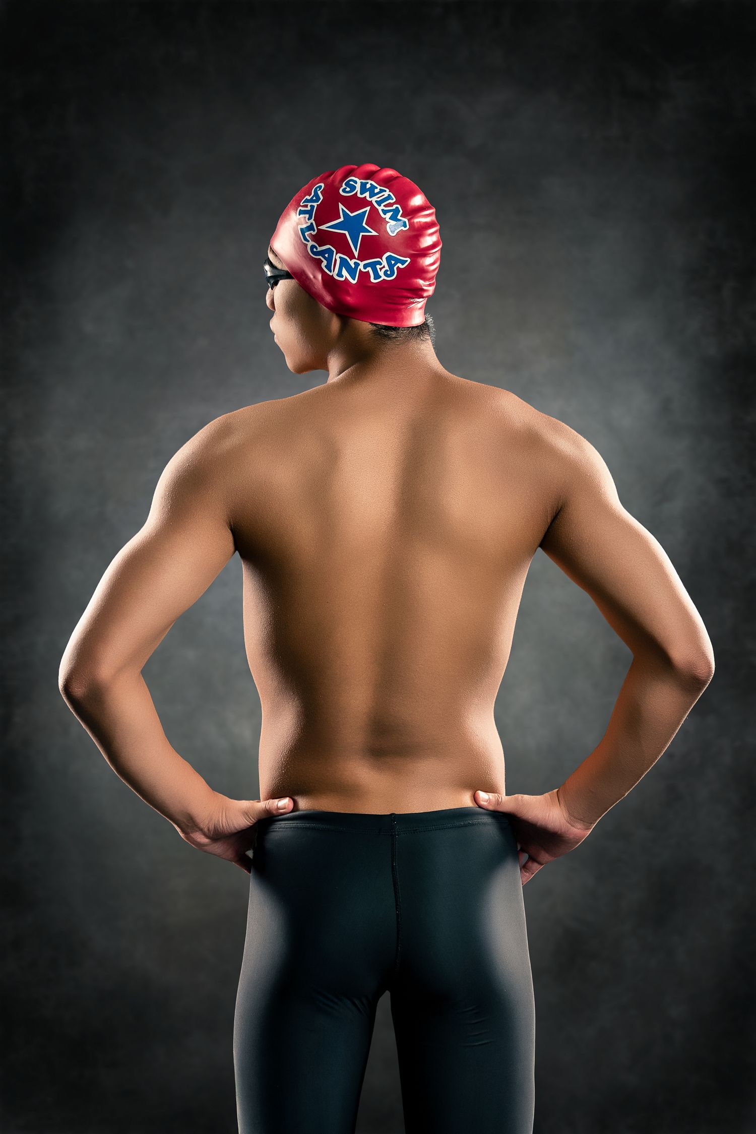 Rear view of swimmer in red Swim Atlanta cap and dark swimwear on gray background.