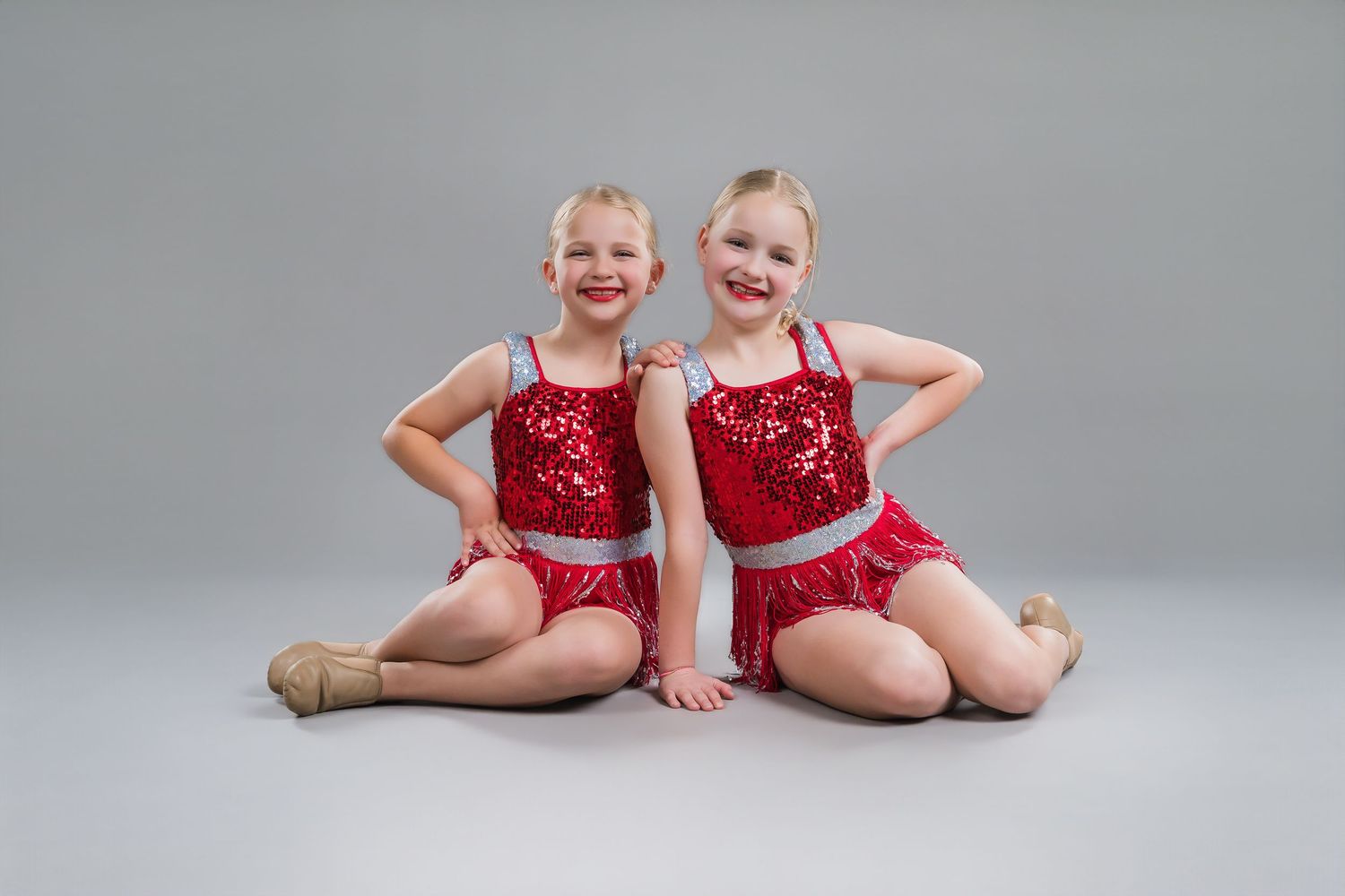 Two young girls in matching red sequin dance costumes sitting together in studio.