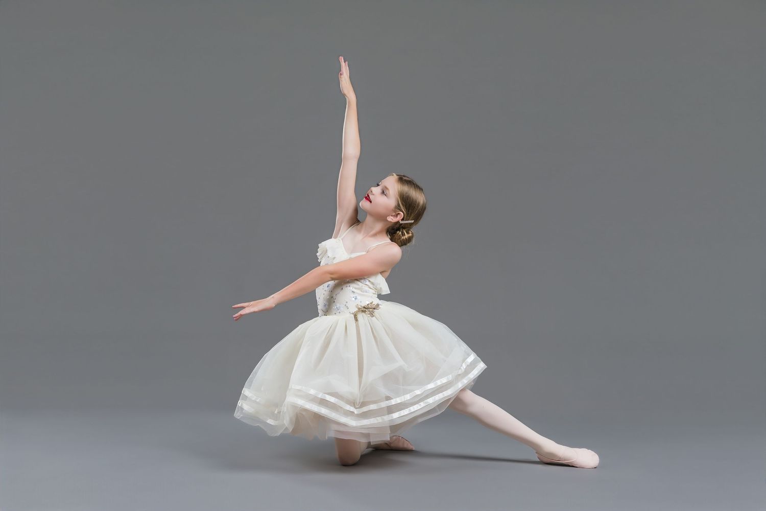 Young ballerina in white tutu dress performing elegant arabesque pose on gray studio background.