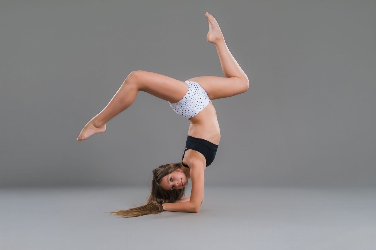 Female gymnast performing headstand with legs split in white polka dot top on gray background.