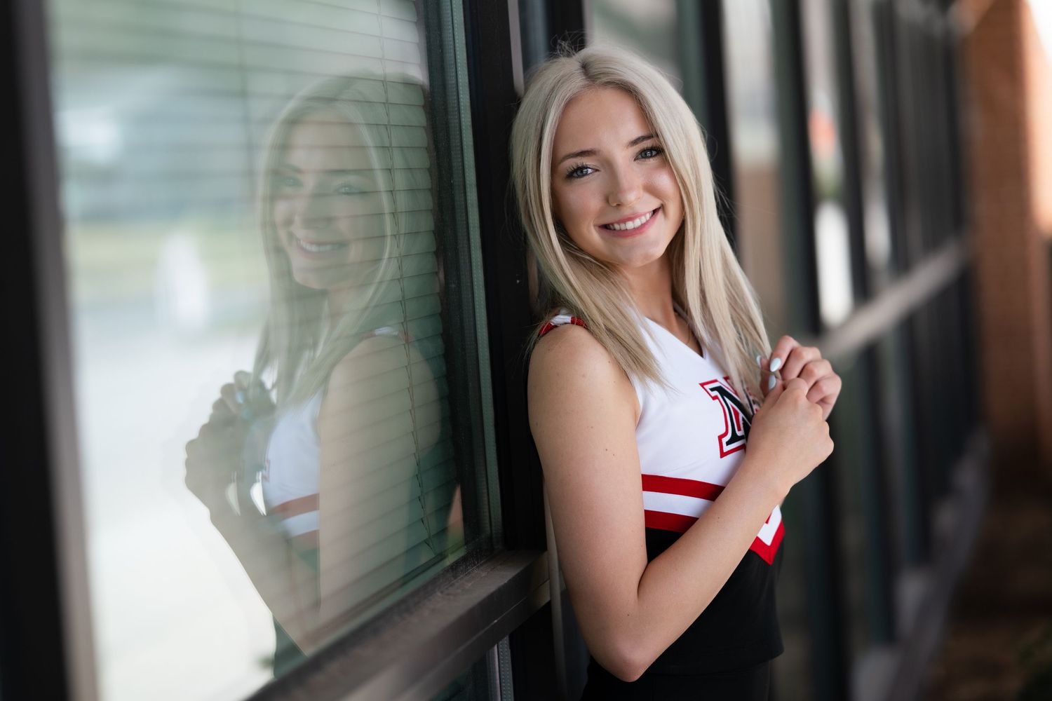 A smiling blonde cheerleader in uniform leans against a window with her reflection visible behind her.