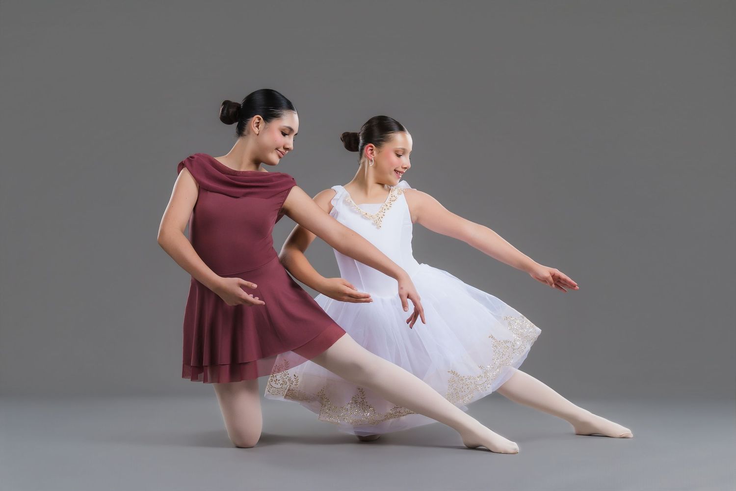 Two female ballet dancers in burgundy and white tutus performing elegant pose on gray background.
