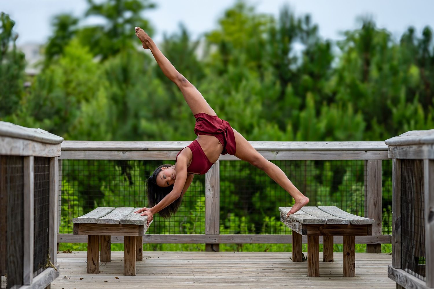 A dancer performs a side split between two benches on a wooden deck surrounded by green trees.
