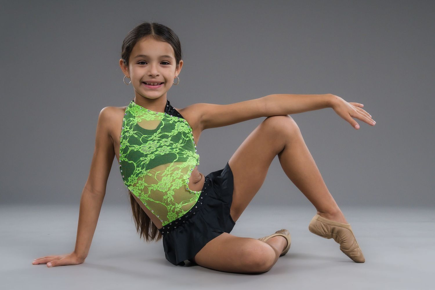 Young girl in green and black dance outfit sitting on studio floor smiling.