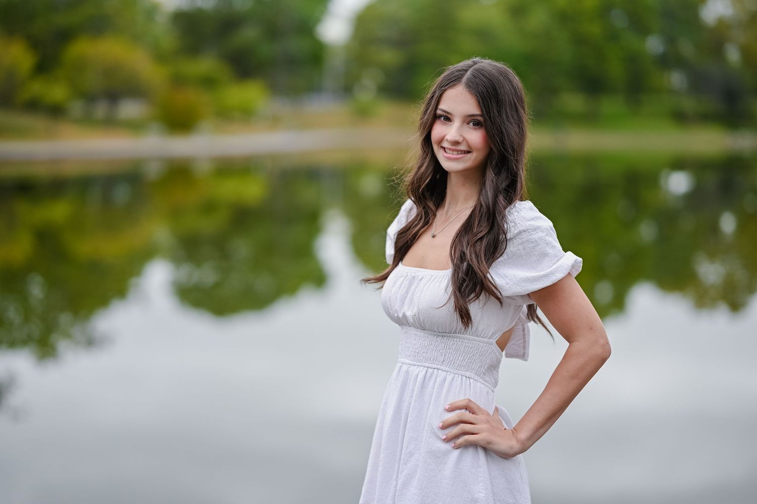 Smiling young woman in white puff-sleeve dress posing by reflective lake in park.