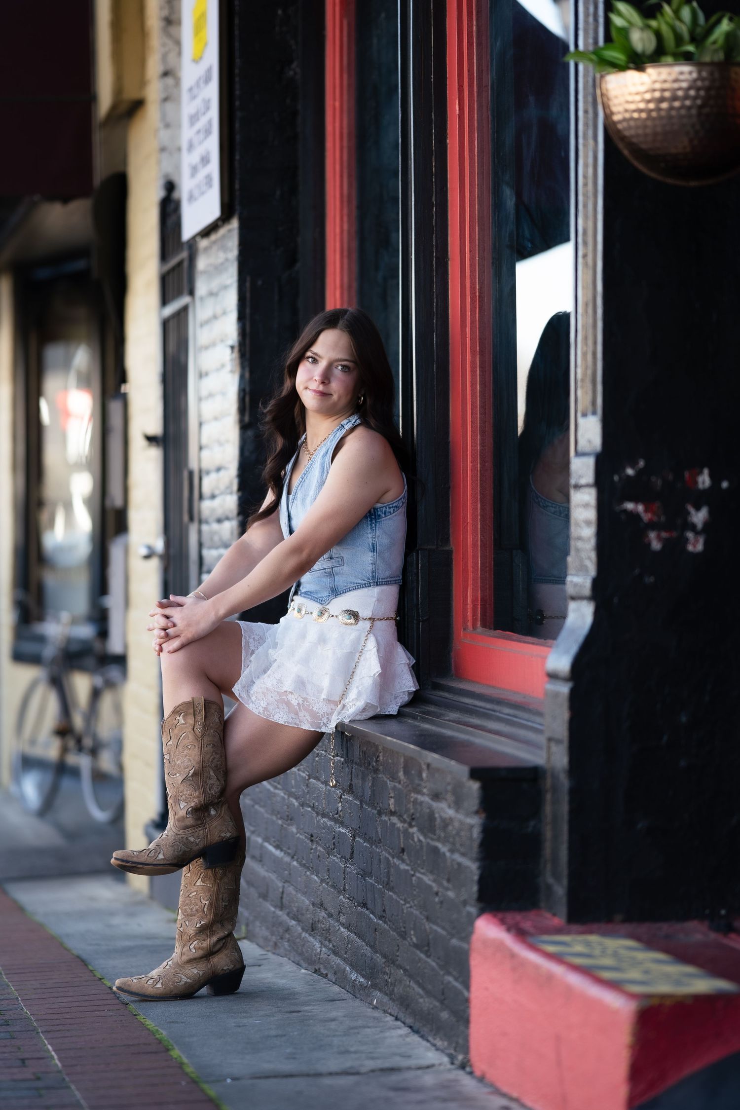 Young woman in denim vest and cowboy boots sitting on storefront ledge on city street.