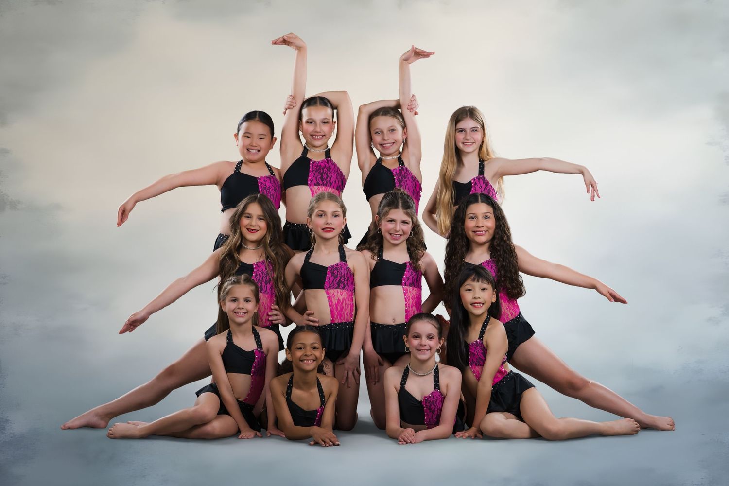 Eleven young dancers in pink and black costumes pose in a tiered group formation for a studio photo.