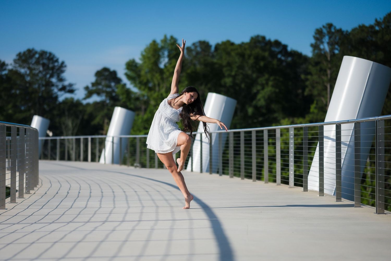 A graceful dancer leaps on an outdoor bridge path, wearing a white dress against a blue sky.