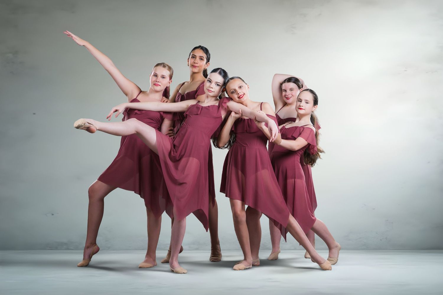 A group of five female dancers pose together in matching burgundy dresses in a studio.