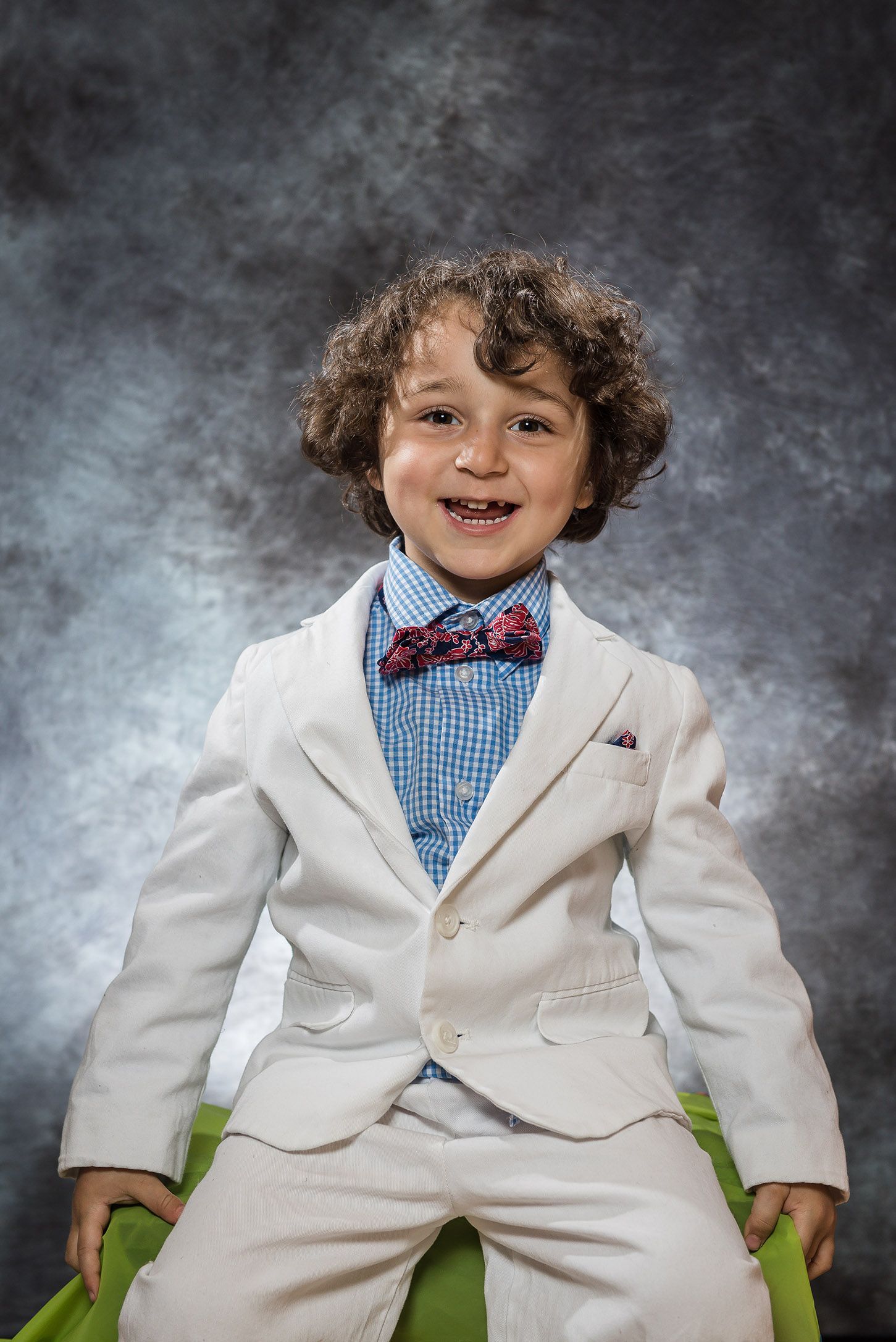 Smiling young boy in white suit with red bow tie and blue checkered shirt posing for portrait photo.