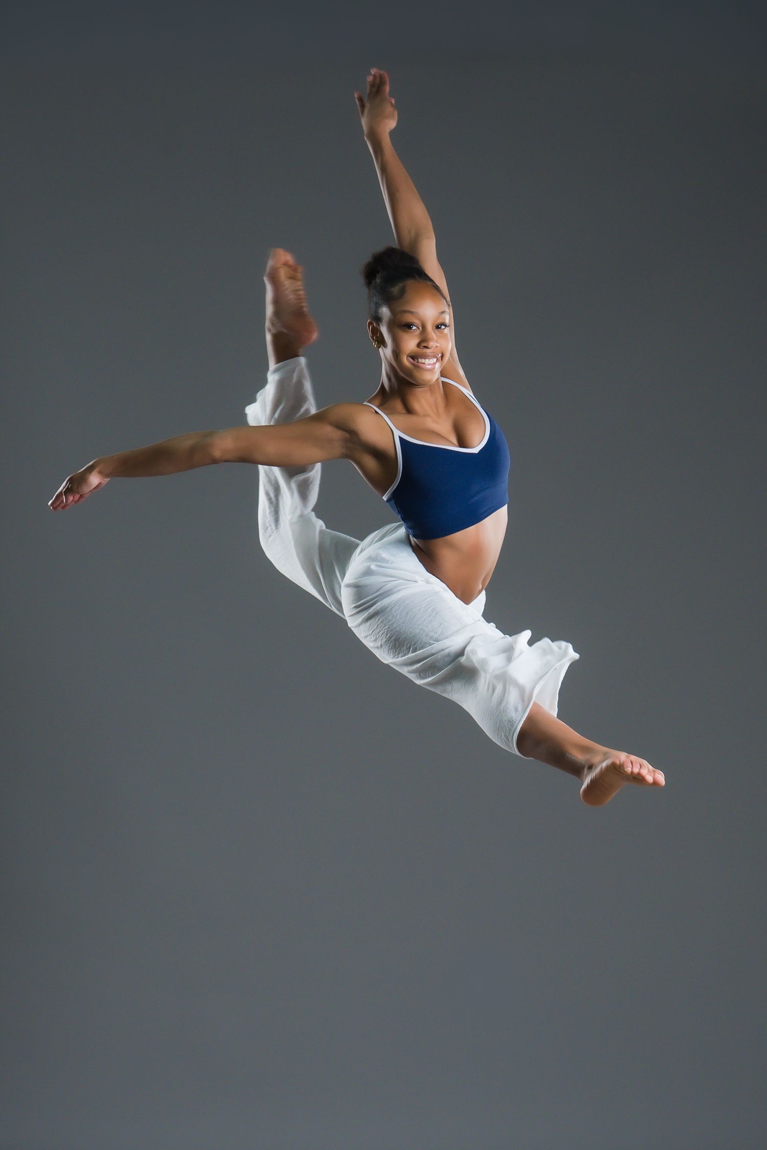 A smiling female dancer leaps gracefully in a blue crop top and white pants against a gray background.