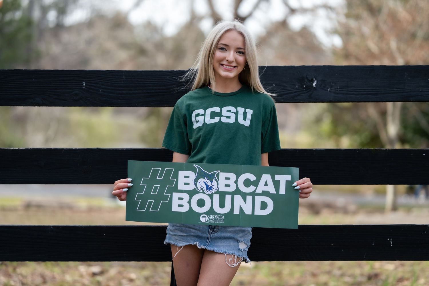 Smiling blonde girl holding GCSU Bobcat Bound sign in front of wooden fence outdoors.
