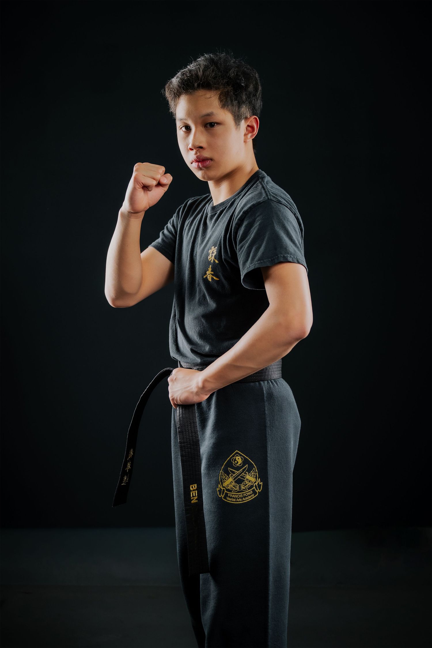 Young martial artist in gray uniform holds black belt, posing in fighting stance against dark background.