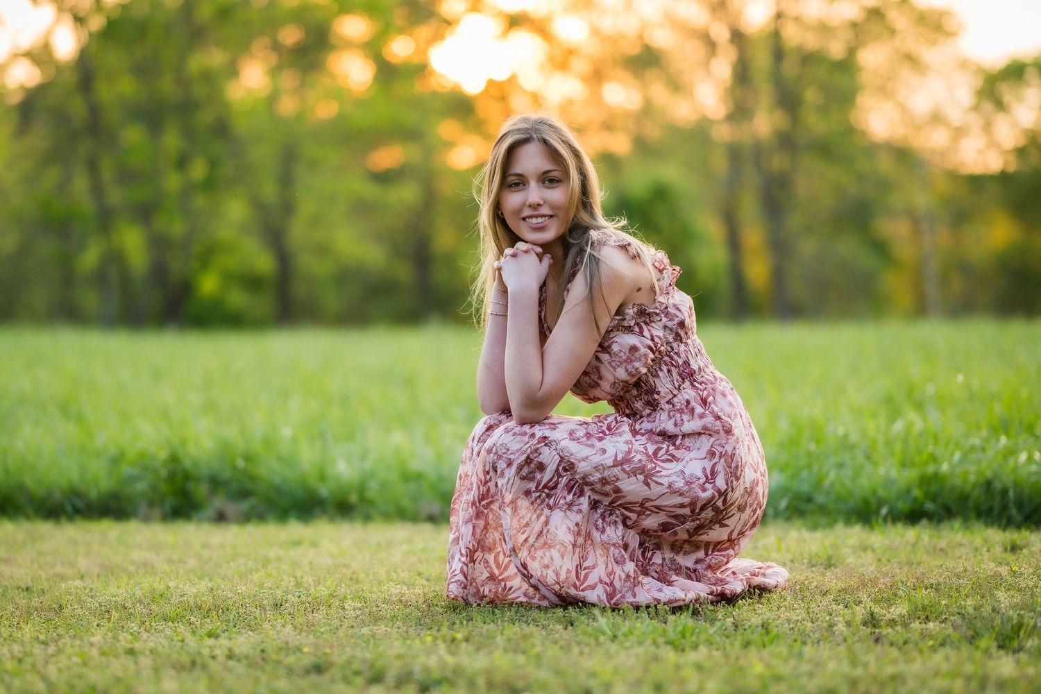 Smiling blonde woman in floral dress sitting on green grass field at golden hour.