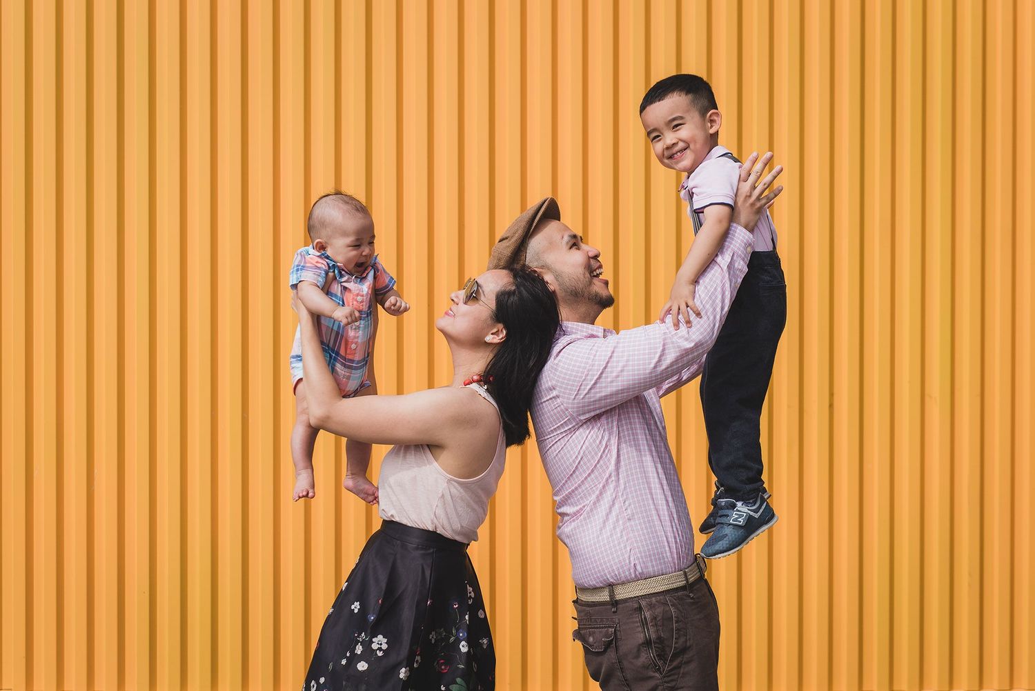 Happy family with two parents lifting their baby and toddler against yellow wall.