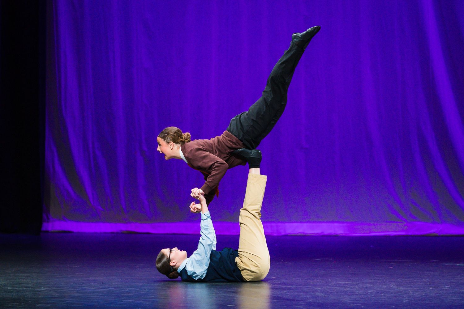Two acrobatic performers executing a balancing act on stage with purple curtain backdrop.