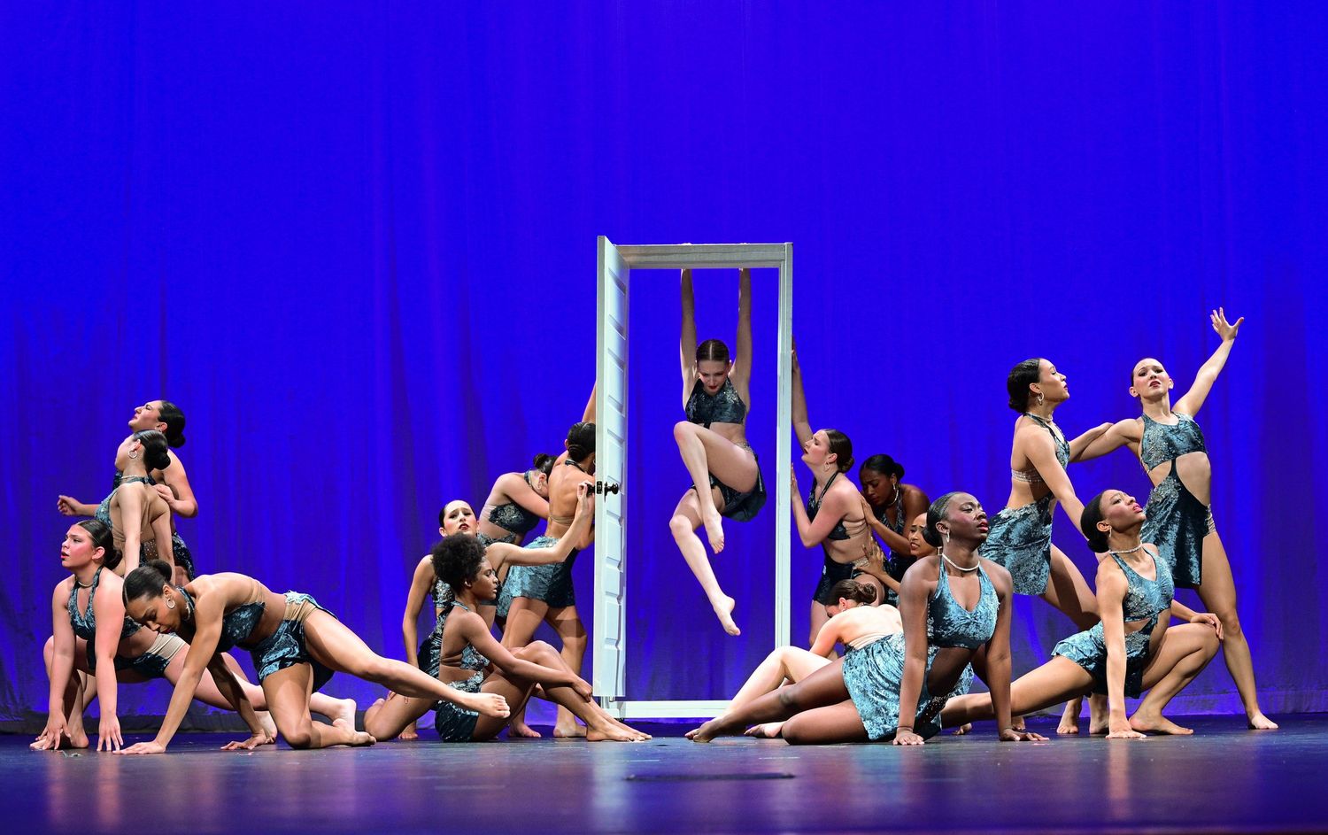 Dancers perform a stunning group routine on a blue-lit stage, with one dancer leaping through a white frame prop.