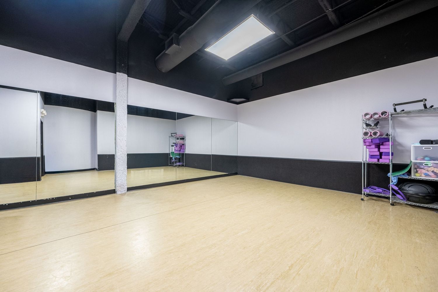 Bright dance studio with hardwood floors, mirrored walls, and a storage rack with yoga mats and equipment.