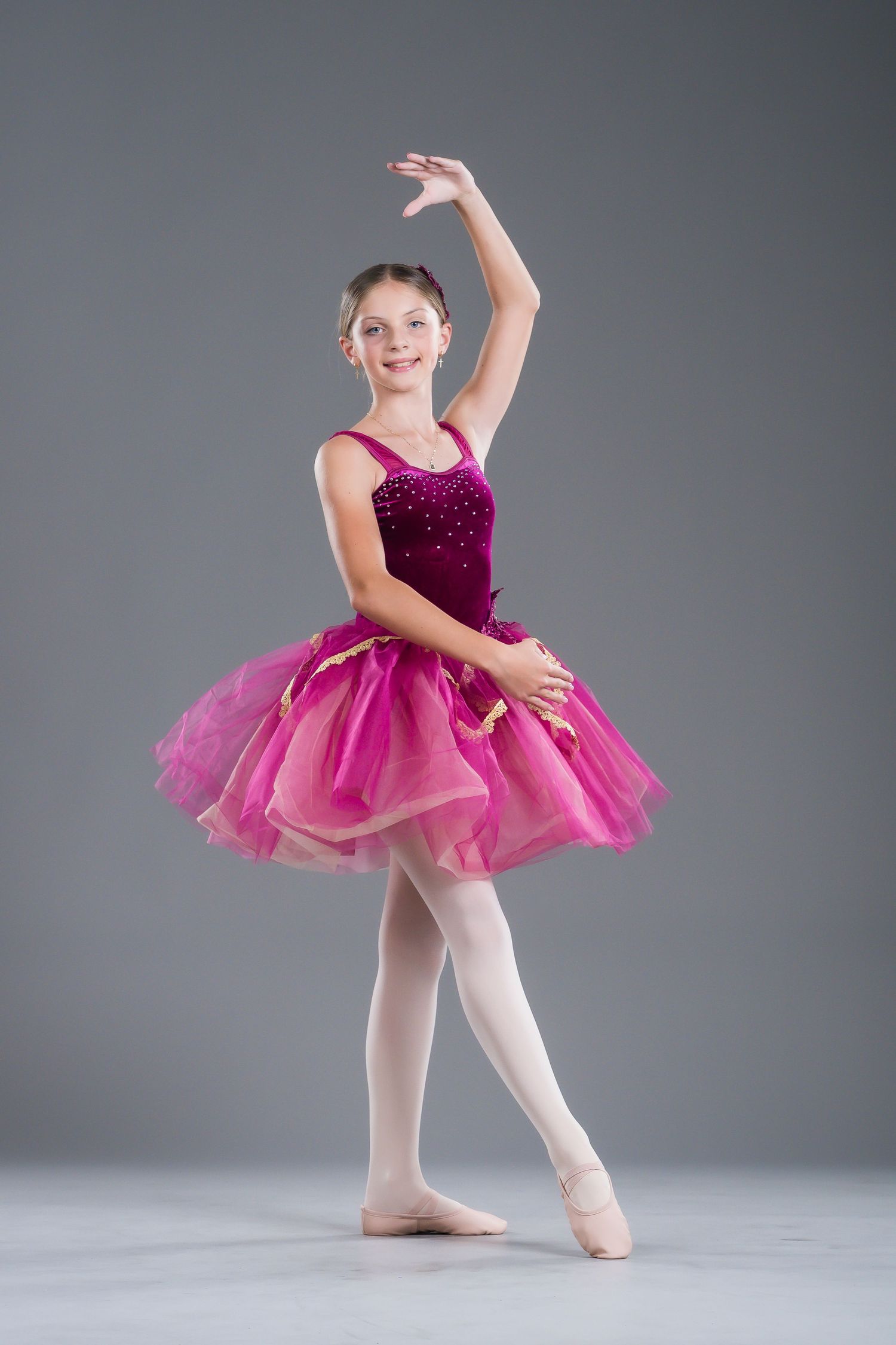 Young ballerina in pink tutu performs graceful pose in dance studio against gray background.