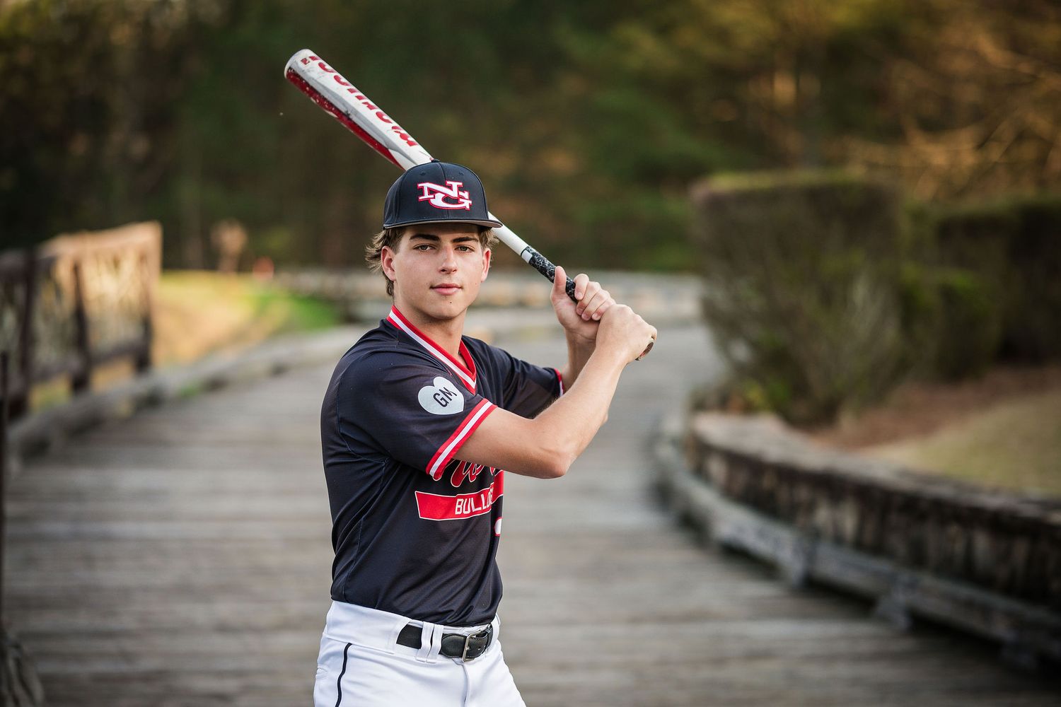 Young baseball player in navy uniform poses with bat over shoulder on wooden bridge outdoors.