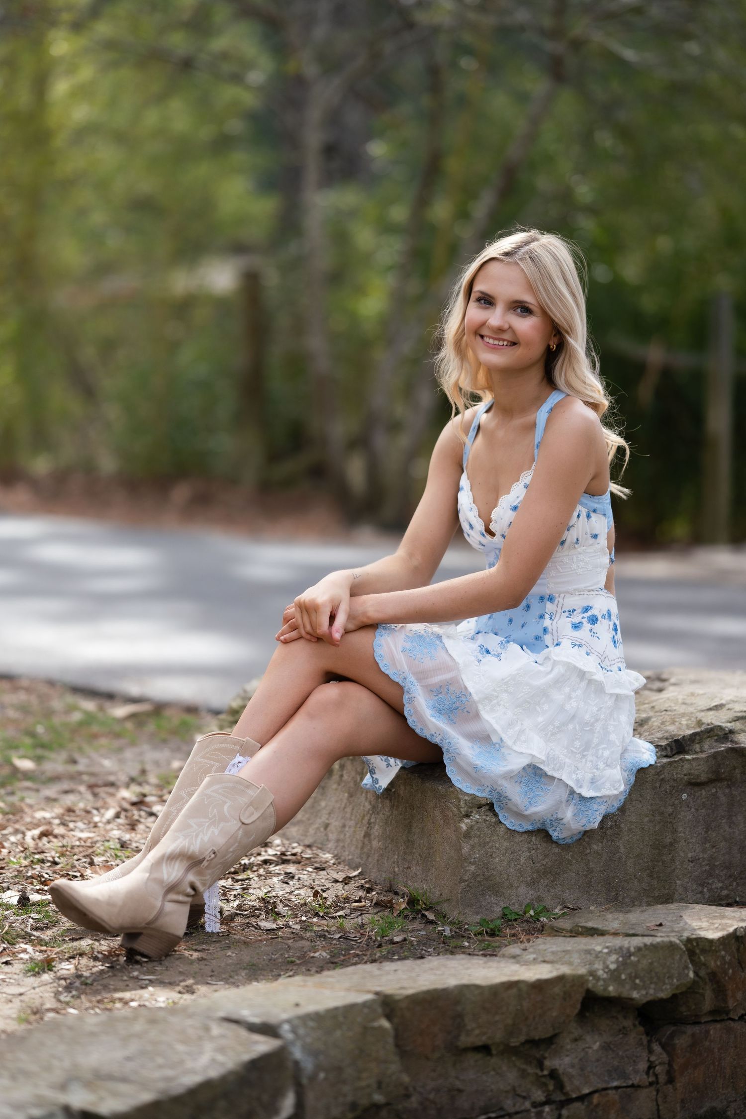 Smiling blonde teen in blue floral dress and cowboy boots sitting on stone steps outdoors.