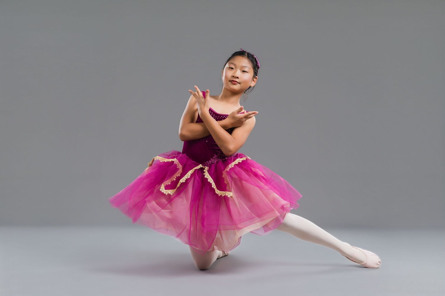Young Asian girl in pink purple ballet tutu performing graceful pose on gray studio background.