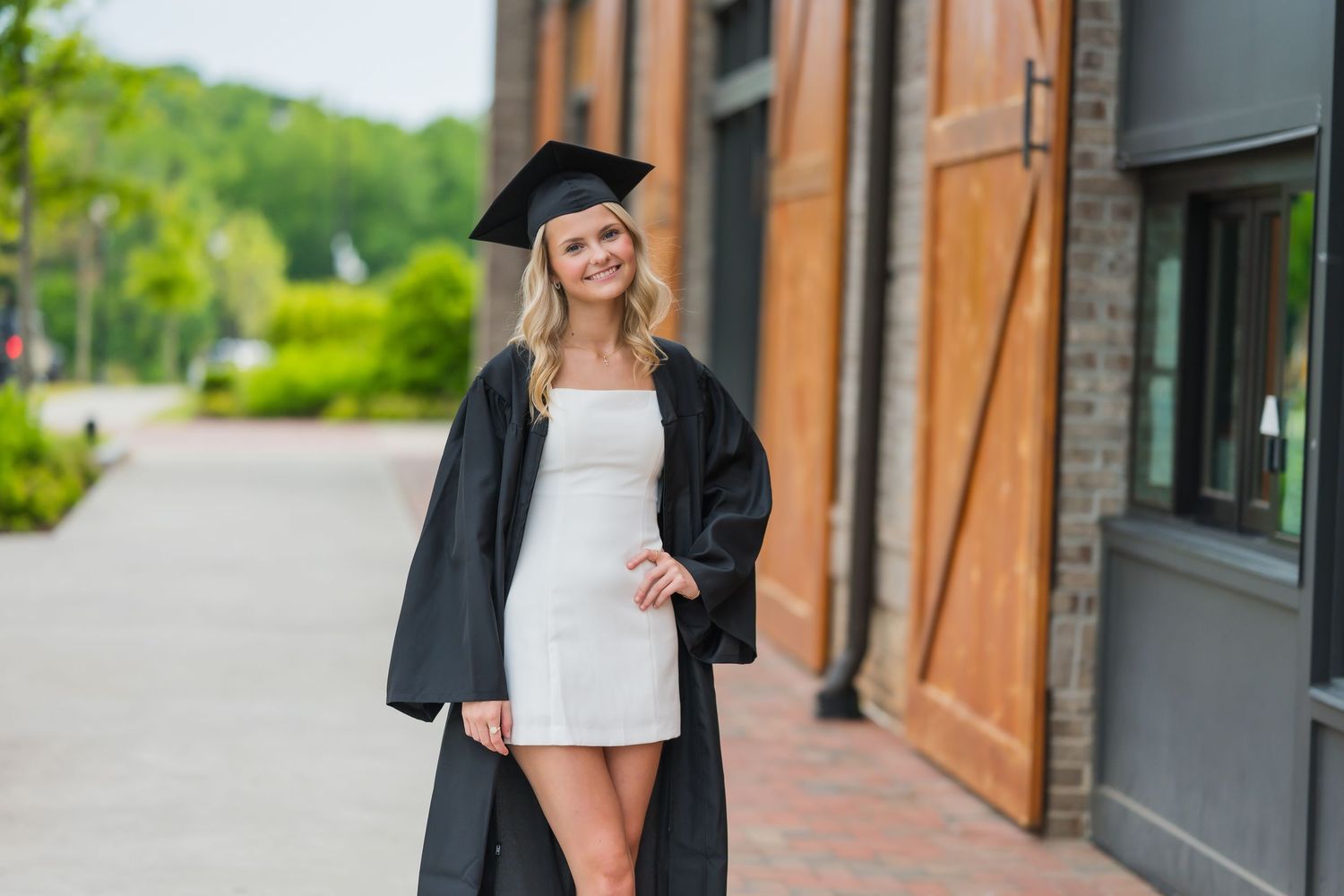 Smiling blonde graduate in black cap and gown wearing white dress posing outdoors.