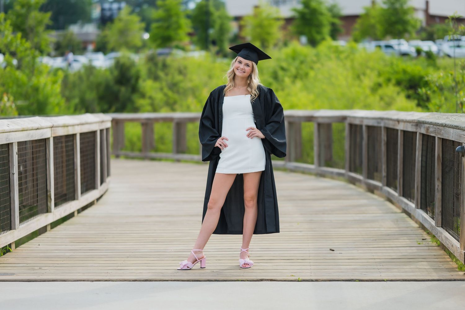A young woman in graduation cap and gown poses on a wooden bridge surrounded by lush greenery.