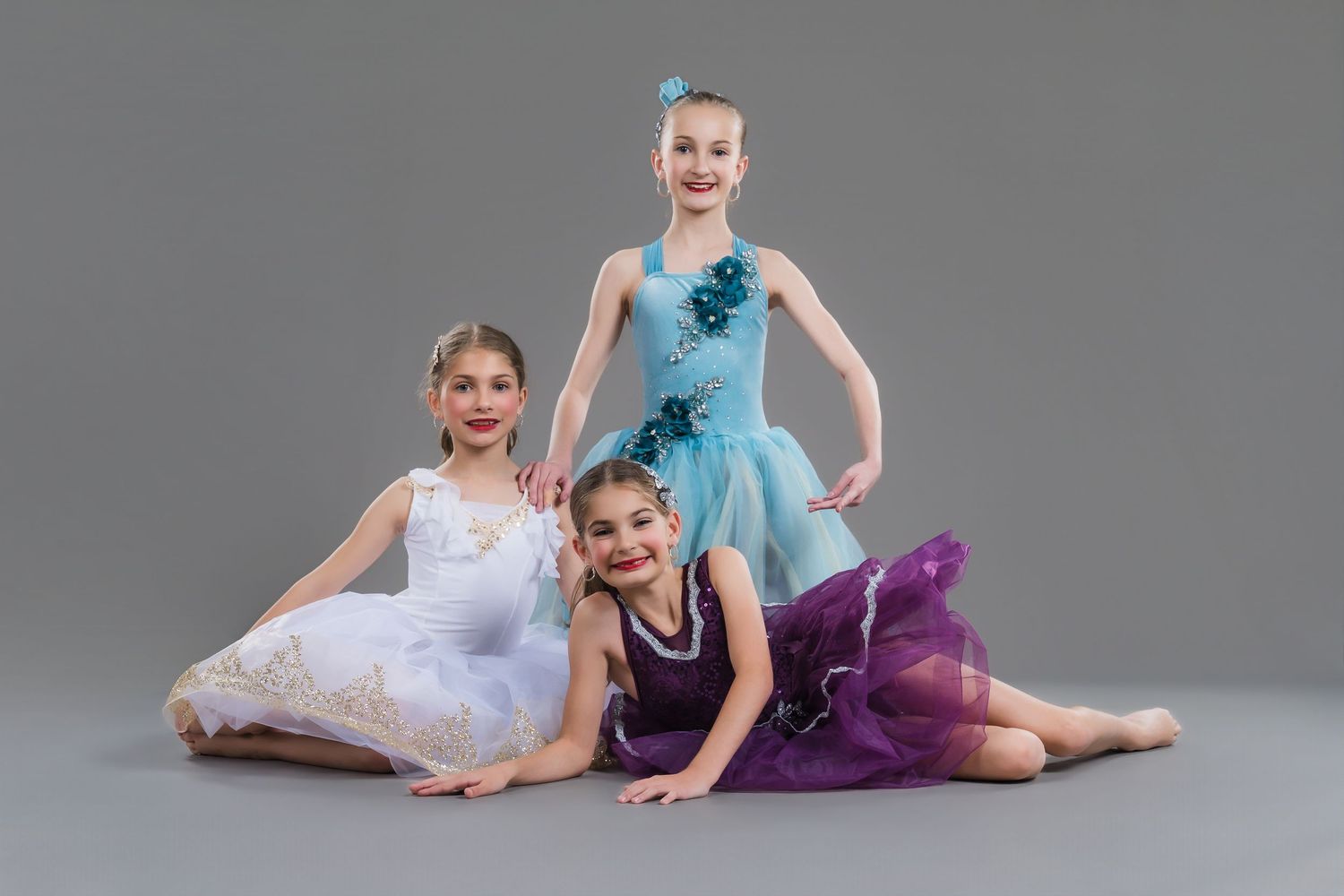 Three young ballet dancers in colorful tutus posing together on gray studio background.
