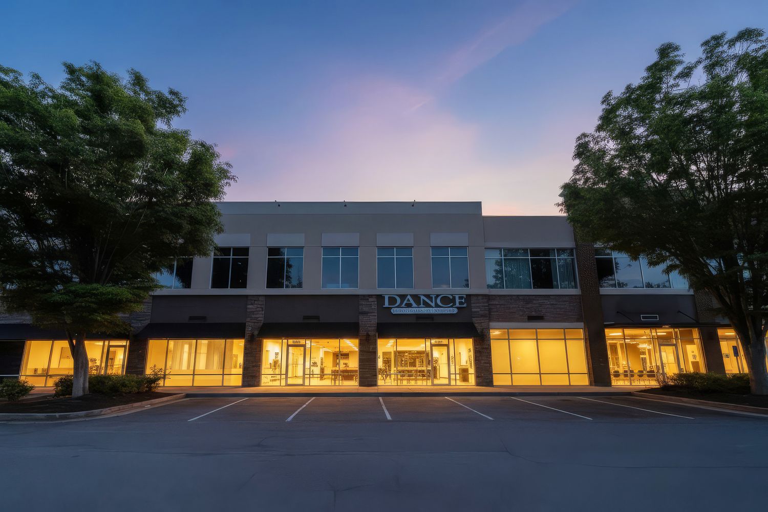 Modern two-story commercial building with illuminated storefronts at dusk, featuring a dance studio and lush trees.