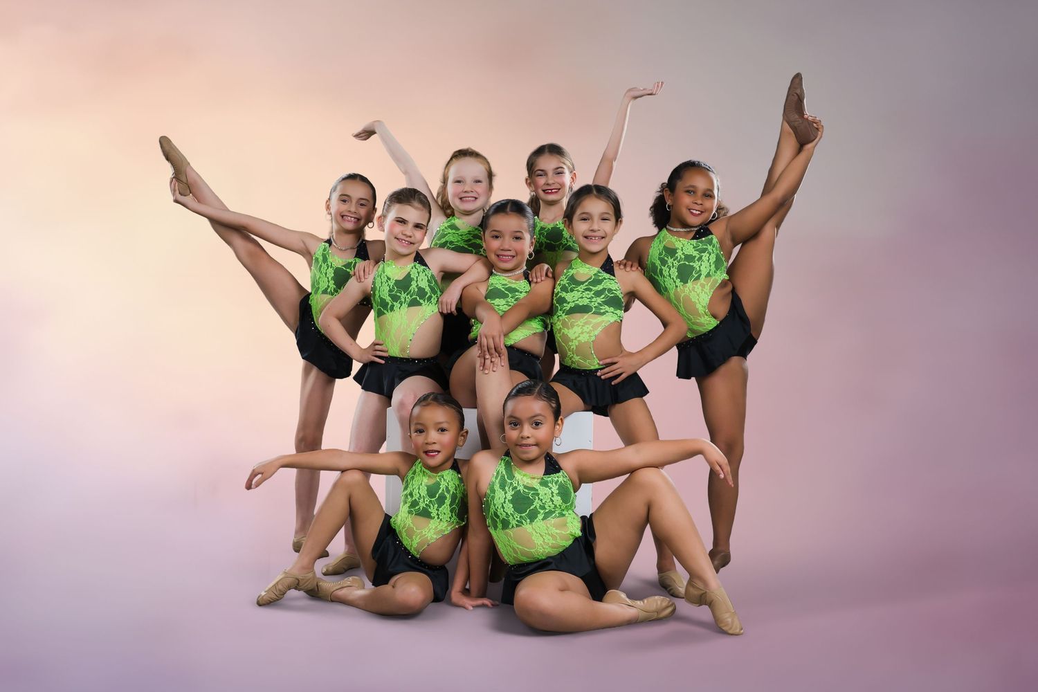 Nine young dancers in green and black costumes pose in a pyramid formation for a studio portrait.