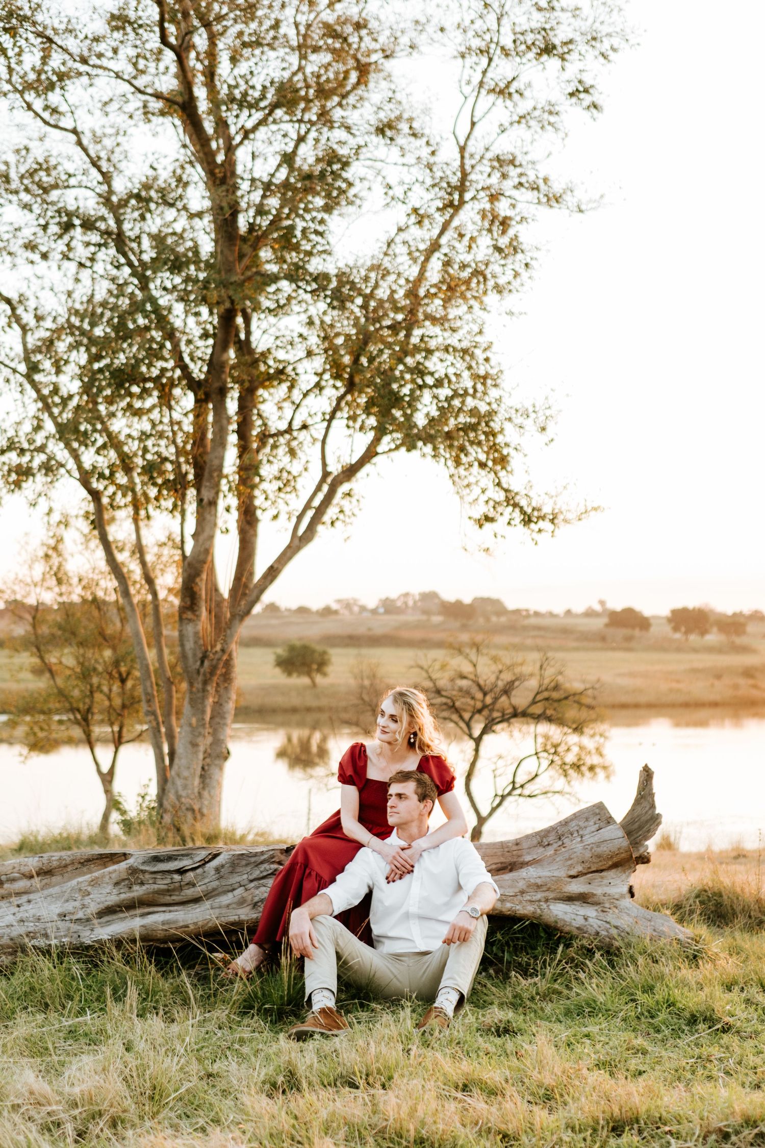 Couple relaxing on a fallen log near a lake at golden hour, woman in red dress sitting behind man.