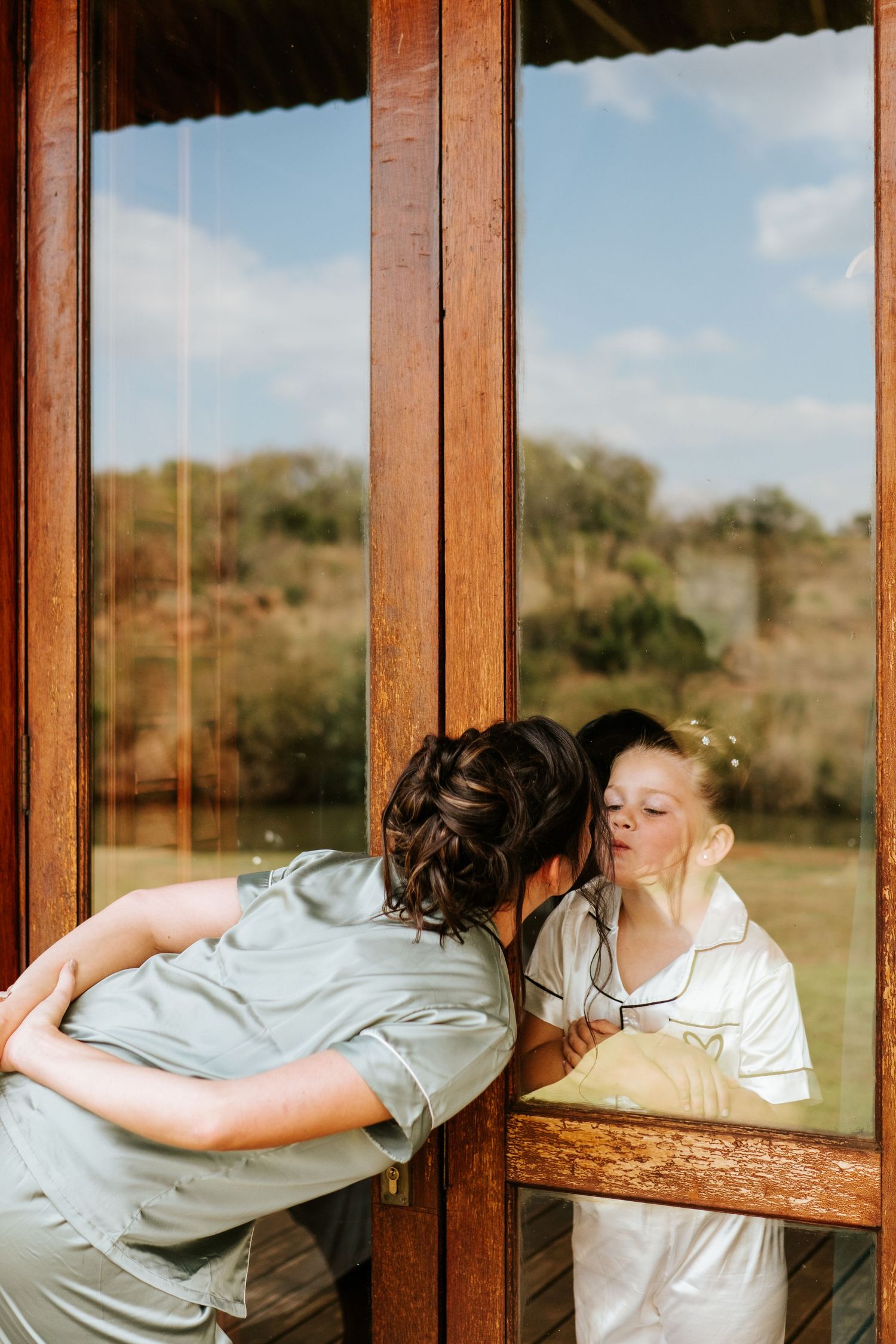 Two women in satin robes sharing a tender moment by rustic wooden glass doors.