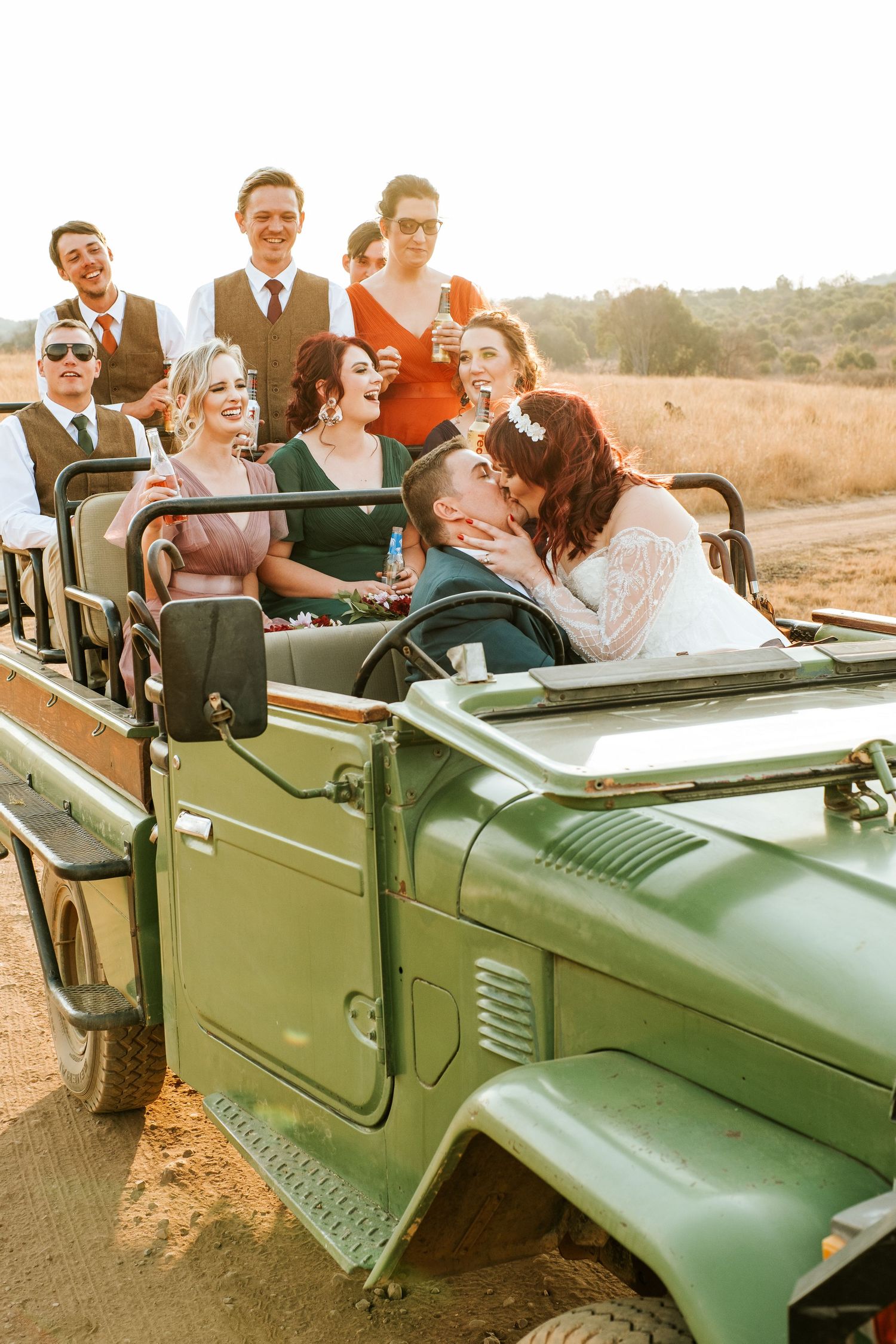 Wedding party laughs and celebrates aboard a vintage green safari jeep at sunset.