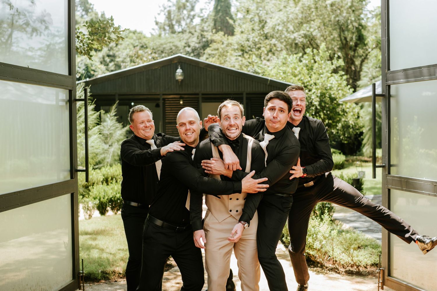 Groom and groomsmen in black suits pose playfully outside a modern glass building.