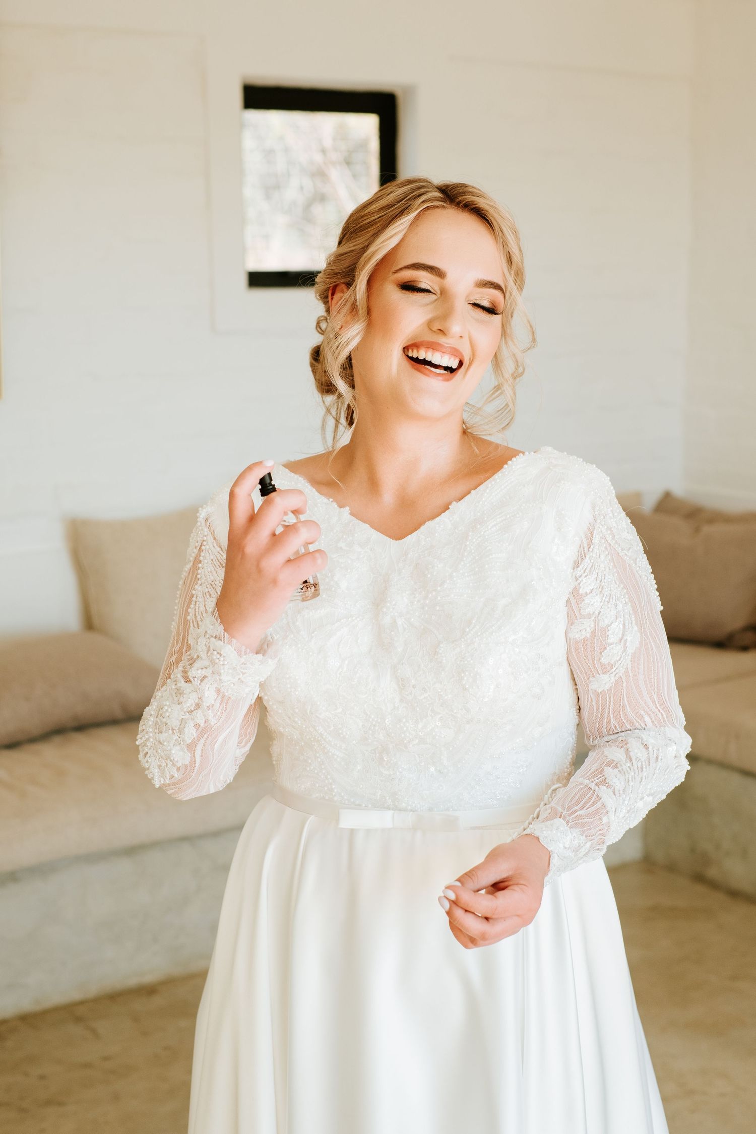 Radiant blonde bride in lace wedding dress laughs joyfully while applying perfume indoors.