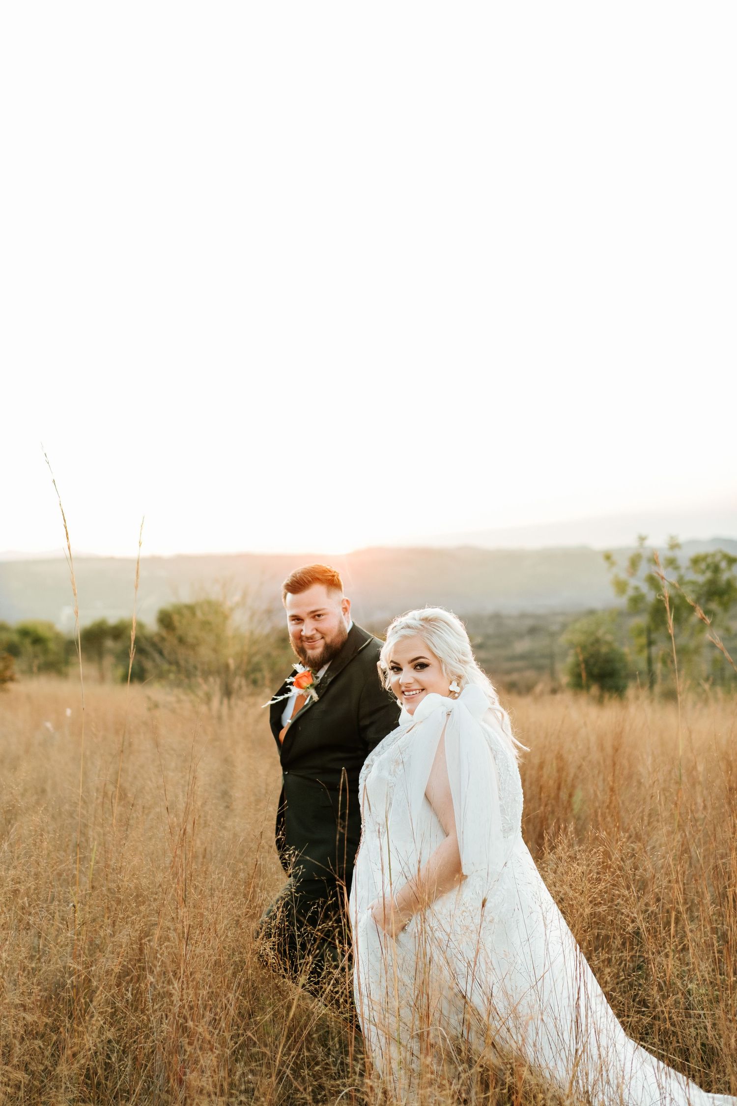 Smiling bride in white lace gown glancing back with groom in black suit in golden sunset field.