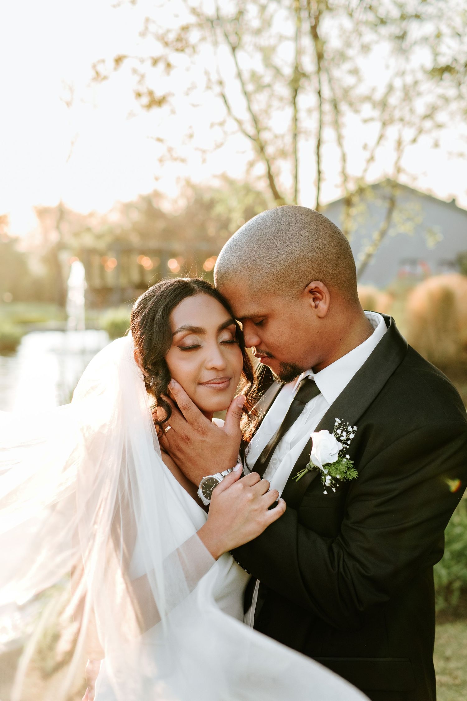 Groom tenderly embracing bride in a veil near a fountain during golden hour wedding portraits.