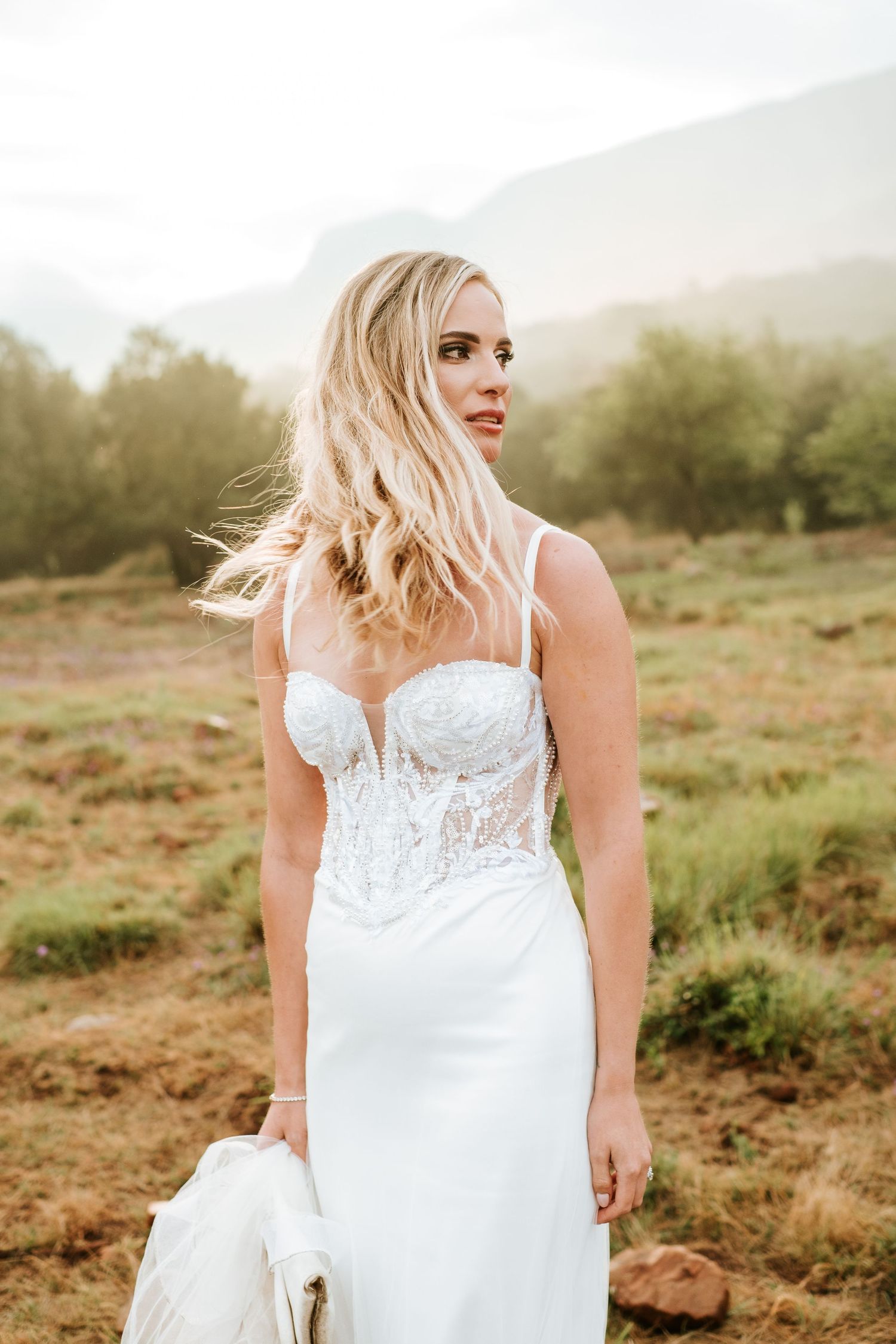 Elegant blonde bride in lace corset gown stands in misty mountain landscape at sunset.