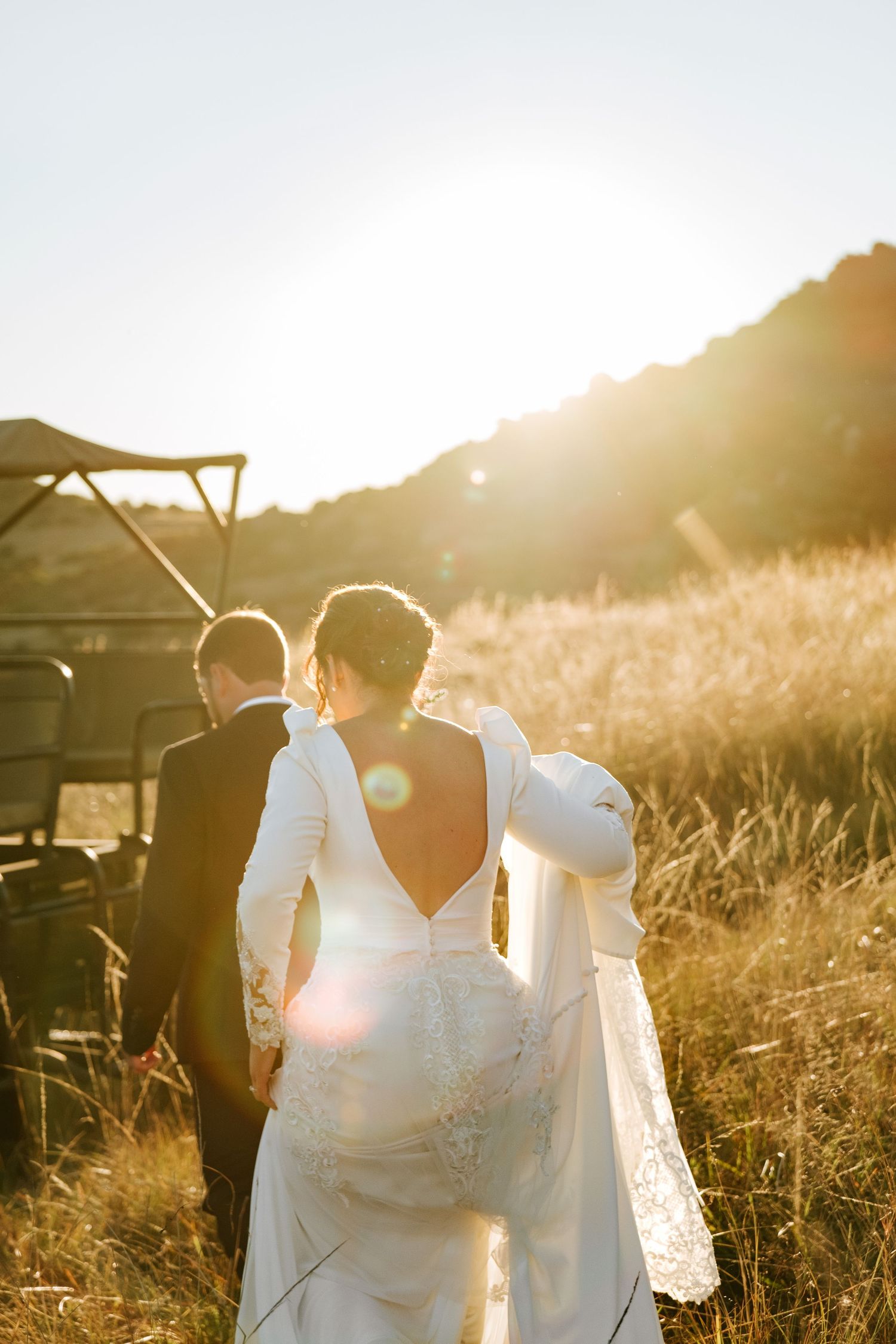 Bride in backless white gown and groom walk toward safari vehicle in glowing sunset field photographed by Laughing Lamp Photo & Film